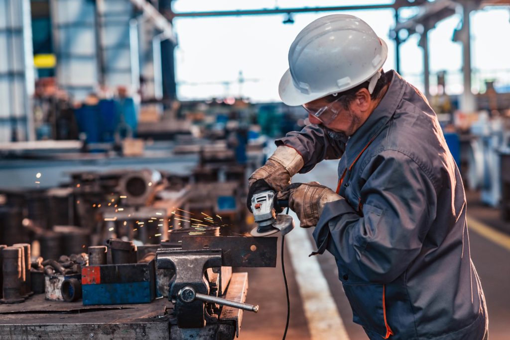 steel worker grinds away at a piece of metal in hard-hat and saftey gear