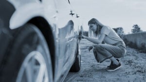Young woman checking air pressure of car tire on local road side while traveling, having troubles with her auto, checking wheel after car breakdown
